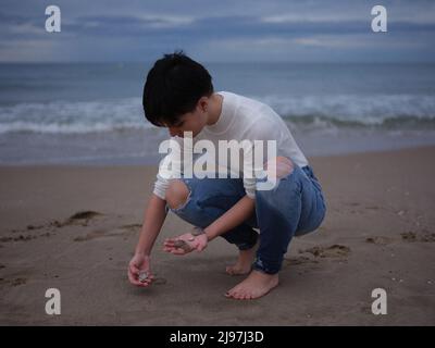 Nicht-binäre Frau, die Muscheln aus dem Sand am Strand aufholt Stockfoto