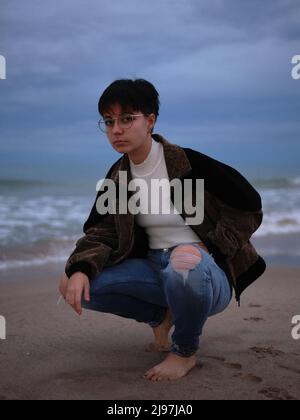 Frau, die barfuß am Strand posiert, trägt lange Hosen und eine Jacke Stockfoto
