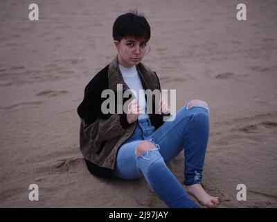 Transgender-Person, die mit einer Jacke und einer Brille auf dem Sand sitzt Stockfoto