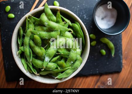 Gedämpftes Edamame, bestreut mit Meersalz auf einem dunklen Steinbrett, Draufsicht Stockfoto