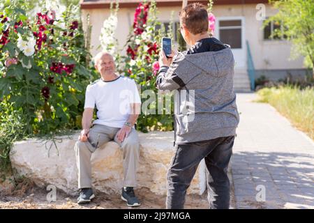 Gesundes Altern. Moderne Senioren sind die besten, die Spaß haben und ein Foto mit einem Smartphone an einem sonnigen Tag in einem Sommergarten machen. Schönes altes Paar im Hinterhof. Selektiver Fokus auf Frau. Stockfoto