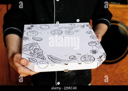 Pizzakoch mit schwarzer Schürze hält frisch gebackene Pizza im Pizzaladen in einer engen Schachtel Stockfoto