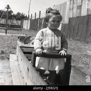 1961, historisches, kleines Mädchen, das auf einem hölzernen, gerahmten Spielplatz, einem Schaukelpferd der damaligen Zeit, Rhyl, Wales, Großbritannien, steht. Ein weiterer klassischer Spielplatzritt der Zeit, der Hexenhut, ist im Hintergrund zu sehen. Stockfoto