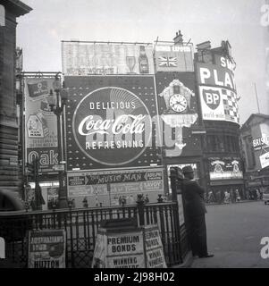 1961, historischer Mann in einem Hut, der am Geländer im Piccadilly Circus, London, England, Großbritannien, steht, mit den berühmten Neon-Werbetafeln, die die Werbetreibenden und Marken des Tages zeigen, darunter Ye Olde Oak Ham, Double Diamond Beer, Players Cigarettes und BP. Das Musical von Meredith Willson, The Music man, ist im Adelphi Theater im West End zu sehen. Stockfoto