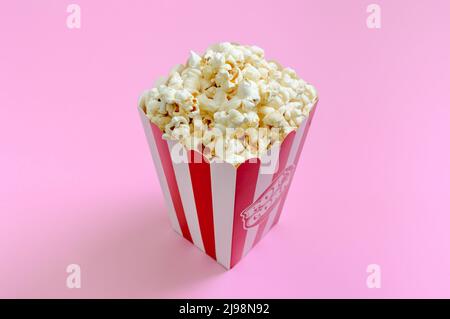 Frisches, knuspriges Popcorn in einem gestreiften Pappbecher auf rosa Hintergrund. Stockfoto