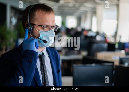 Ein Mann mit Latexhandschuhen und einer medizinischen Maske spricht über das Headset mit einem Kunden. Ein männlicher Büroleiter nimmt Anrufe entgegen. Arbeiten während des Coronavirus Stockfoto