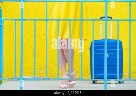 Weibliche Beine in hochhackigen Schuhen mit einem blauen Koffer für Geländerschuhe auf gelbem Hintergrund. Stockfoto