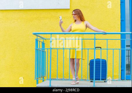 Schöne rothaarige Frau steht mit einem blauen Koffer und nutzt ein Smartphone auf gelbem Hintergrund. Stockfoto