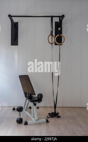 Zimmer ohne Leute mit Ringen und einer Pull-up-Bar im Fitnessraum. Hanteln neben einer Bank. Weißer Hintergrund und Holzboden. Gesunder Lebensstil Stockfoto