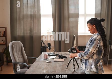 Horizontale Aufnahme einer stilvollen jungen Frau mit afro-Zöpfen in legerem Outfit, die im Loft-Raum am Schreibtisch steht und die Kameraeinstellungen überprüft, um Videos aufzunehmen Stockfoto