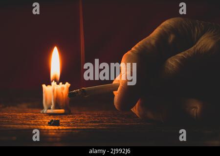 Die alte Frau hält eine Zigarette bei Kerzenlicht im Dunkeln zu Hause am Tisch, raucht zu Hause im Dunkeln, Nikotin und eine schlechte Angewohnheit, Zigaretten und Kn Stockfoto