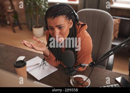 Porträt einer jungen Influencerin mit Kopfhörern, die am Schreibtisch sitzt und sich verwirrt fühlt, als sie seltsame Kommentare zu ihrem Inhalt liest Stockfoto
