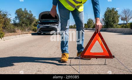 Auto mit Problemen und Mann legte ein rotes Dreieck, um andere Verkehrsteilnehmer zu warnen. Männlicher Fahrer, der in der Nähe eines kaputten Autos mit offener Motorhaube steht und auf Hilfe wartet. Stockfoto