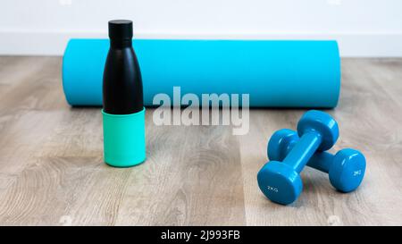 Trainieren Sie mit zwei blauen Kurzhanteln mit moderner Öko-Wasserflasche und Matte, um sich auf das Yoga-Sporttraining im Haus vorzubereiten. Meditieren und Fitnesstraining Stockfoto