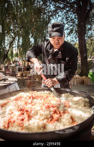 Ein männlicher Koch kocht Pilaf in einem großen Kessel auf der Straße. Stockfoto