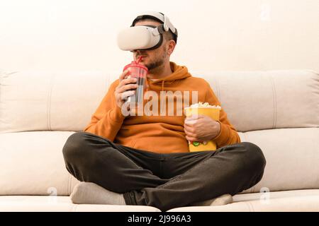 Mann, der einen Film in seiner Virtual-Reality-Brille sieht, trinkt Limonade und hält Popcorn, während er auf der Couch sitzt Stockfoto