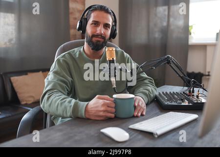 Mittleres Porträt eines hübschen jungen Bloggers aus dem Nahen Osten, der Kopfhörer trägt und eine Tasse Tee am Schreibtisch hält und die Kamera anschaut Stockfoto