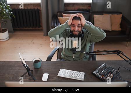 Horizontaler Weitwinkelaufnahme eines modernen bärtigen jungen Influencers aus dem Nahen Osten, der in legerer Kleidung am Schreibtisch im Loftzimmer sitzt und sich aufregt Stockfoto