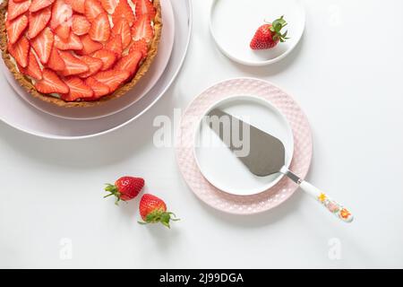 Köstliche Tarte mit Pudercreme und Erdbeeren auf einem weißen Tisch. Teller und Teigschaber zum Auftragen des Teigs. Blick von oben. Stockfoto