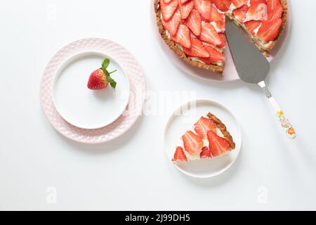 Köstliche Tarte mit Pudercreme und Erdbeeren auf einem weißen Tisch. Teller und Teigschaber zum Auftragen des Teigs. Stück geriebenen auf einem Teller. Blick von einem Stockfoto