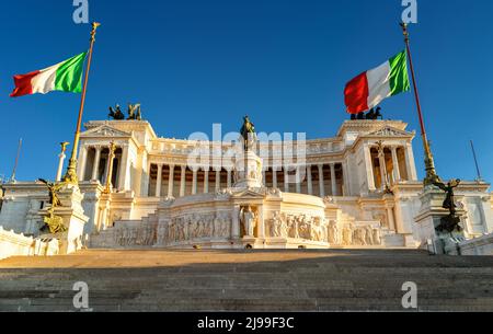 Vittoriano-Gebäude mit italienischen Flaggen auf dem Venedigplatz bei Sonnenuntergang, Rom, Italien. Es ist das Wahrzeichen Roms. Sonniger Blick auf die klassische Architektur im Capitol Stockfoto