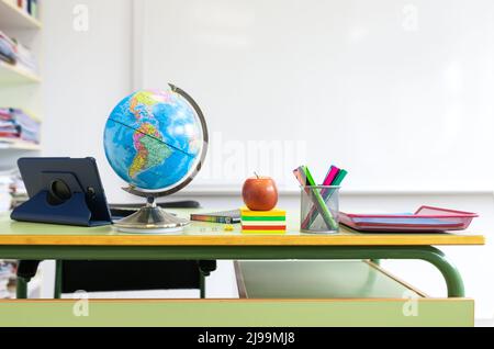 Detail eines Schulklassenzimmers. Der Tisch des Lehrers mit Objekten darauf. Tablet und Tastatur, Weltkugel, Notebook, Büroklammer und Binderclip, Haftnotizen Stockfoto