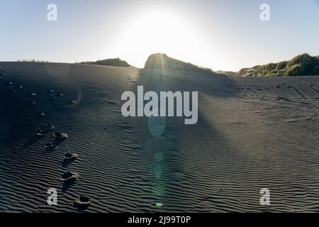 Wellen und Bahnen sich durch den schwarzen Sand der sanft geschwungenen Castlecliff-Dünen, während Sonnenuntergänge hinter ihnen einen Lichteffekt erzeugen, Whanganui, Neuseeland. Stockfoto