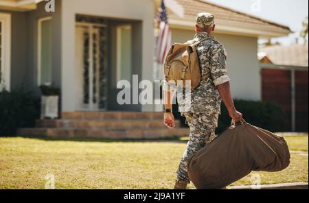 Rückansicht eines patriotischen jungen Soldaten, der mit seinem Gepäck auf sein Haus zuläuft. Mutiger amerikanischer Militärangehöriger, der nach dem Verrichten seines CO nach Hause zurückkehrt Stockfoto