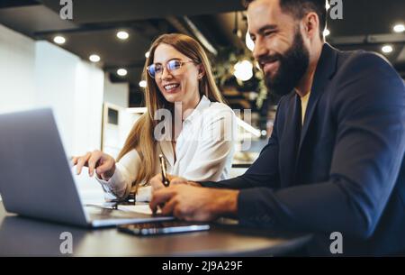 Fröhliche Geschäftsleute, die in einem Büro einen Laptop benutzen. Glückliche junge Unternehmer lächeln, während sie in einem modernen Arbeitsbereich zusammenarbeiten. Zwei junge Geschäftsleute Stockfoto