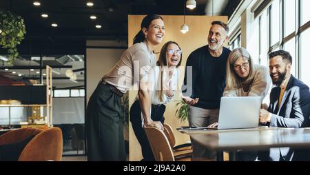 Fröhliche Geschäftsleute, die lachen, während sie an einem neuen Projekt in einem Büro mitarbeiten. Eine Gruppe verschiedener Geschäftsleute, die während der Zusammenarbeit einen Laptop verwenden Stockfoto