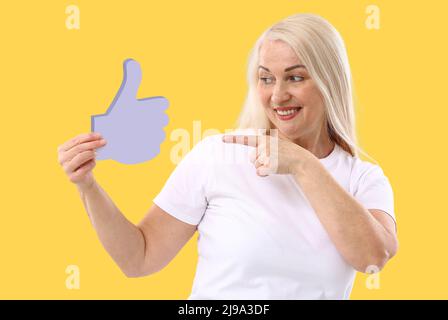 Reife Frau mit Daumen-up-Symbol auf gelbem Hintergrund Stockfoto