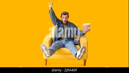 Lustiger junger Mann mit Eimer Popcorn, der auf gelbem Hintergrund in einem Sessel sitzt Stockfoto