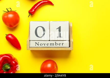 Holzwürfelkalender mit Datum NOVEMBER 1 und Gemüse auf gelbem Hintergrund, Nahaufnahme. Konzept des Welt-Vegan-Tages Stockfoto