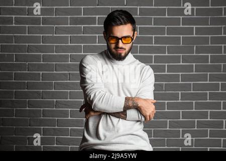 Junger bärtiger Mann mit gekreuzten Händen und Sonnenbrille auf grauem Backsteinwand-Hintergrund Stockfoto