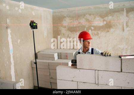 Maurer mit einem Gummihammer zum Klopfen und Ausgleichen der Betonblockwand. Der Auftragnehmer verwendet präzise Wasserblase und Laserwaage für Mauerwerk Stockfoto