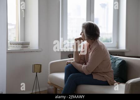 Traurige einsame, reife, grauhaarige Dame, die vom Fenster weg schaute Stockfoto