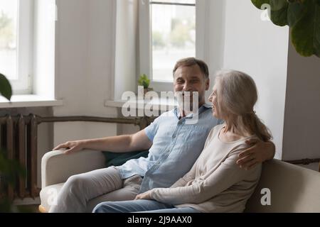 Fröhliches, fröhliches, liebevolles, älteres Paar, das redet, lacht und sich auf der Couch umarmt Stockfoto