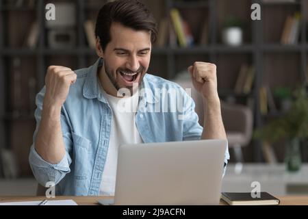 Aufgeregter junger Geschäftsmann, der von zu Hause aus arbeitet, hebt die Fäuste und schreit yeah Stockfoto