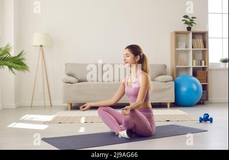 Ruhige und entspannte junge Sportlerin, die morgens zu Hause im Raum mit Sonnenlicht meditiert. Stockfoto