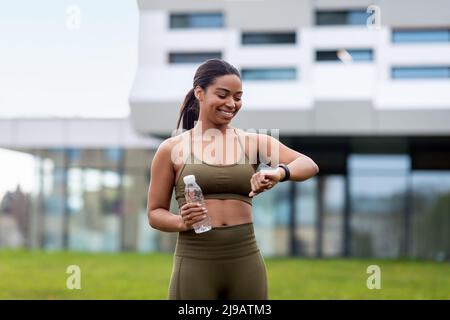 Porträt einer sportlichen jungen schwarzen Frau mit einer Flasche Wasser, die ihre Smartwatch oder ihren Fitness-Tracker im Freien überprüft Stockfoto