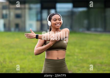 Fröhliche junge schwarze Frau, die in Kopfhörern Musik hört, ihren Arm streckt und im Stadtpark Sport macht Stockfoto