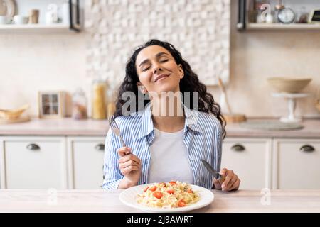 Aufgeregte Dame genießt köstliche hausgemachte Pasta, leckeres Mittagessen mit geschlossenen Augen, während sie an einem Tisch in der Küche sitzt Stockfoto