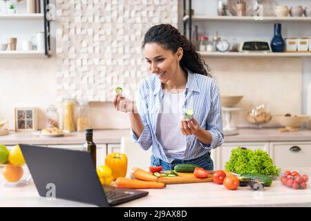 Glückliche lateinerin, die auf dem Laptop ein Abendessen kocht und Videoanrufe macht, mit der Webcam spricht und in ihrer Wohnung in der Küche steht Stockfoto