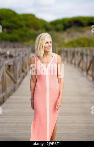 Schöne reife Frau, die einen Holzweg in der Nähe des Strandes entlang geht, in einem schönen orangefarbenen Kleid. Stockfoto