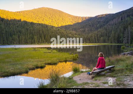Eine europäische Frau, die bei Sonnenuntergang auf einer Holzbank am schönen See kleiner Arbersee sitzt. Blick auf den Großen Arber mit seinen zwei Türmen. Bayerische Front Stockfoto