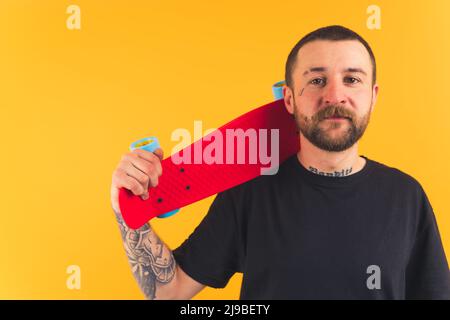 Mittlere Nahaufnahme von jungen kaukasischen Mann mit einem Tattoo auf seinem Gesicht hält roten Skateboard auf seiner Schulter gelben Hintergrund Studio Schuss Kopie Raum . Hochwertige Fotos Stockfoto