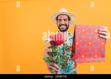 Lateinischer bärtiger Mann, der ein Geschenk gab und eine Rose auf der Kamera über orangefarbenem Hintergrund, isoliert vom Studio. Hochwertige Fotos Stockfoto