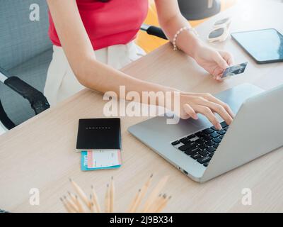 Die Hand der jungen Frau hält Kreditkarte und Reisepass, während sie den Laptop für die Flugbuchung benutzt, das Hotelzimmer für die Sommerreise auf dem Tisch, bereit für die Reise Stockfoto