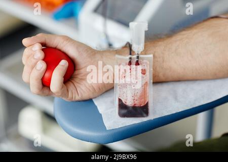 Nahaufnahme der männlichen Hand, die den Stressball drückt, während sie Blut im medizinischen Freiwilligenzentrum spendet, konzentrieren Sie sich auf den Blutbeutel im Vordergrund, Platz kopieren Stockfoto