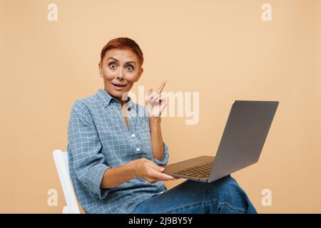 Senior überraschte Frau beim Arbeiten mit einem Laptop, der über einer beigen Wand isoliert war Stockfoto